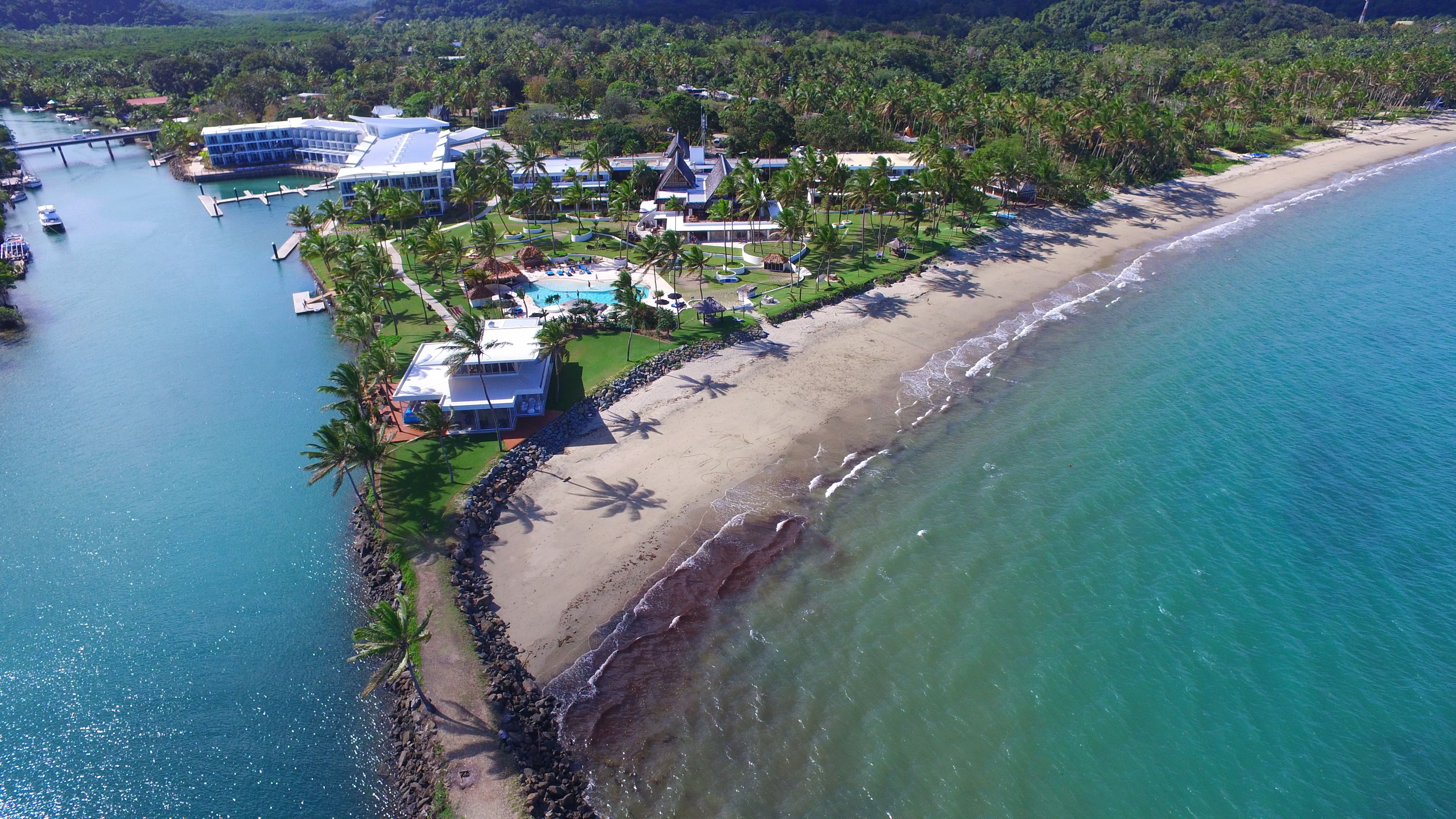 An aerial view of a coastal resort with a beach, a swimming pool, and a bridge connecting to a dock.