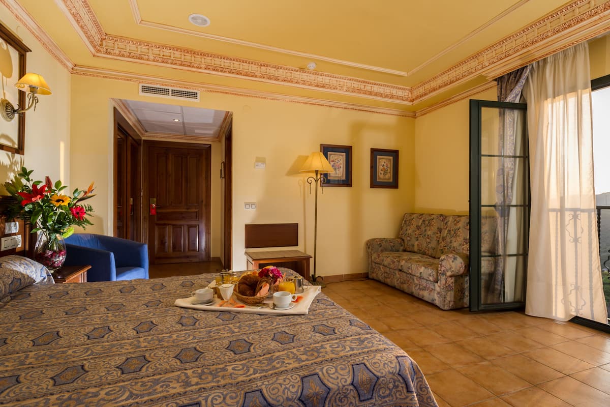 Ein Hotelzimmer mit einem großen Bett, einer Sitzecke mit einer Couch, einem Esstisch mit Frühstück und einem Balkon mit Vorhängen.