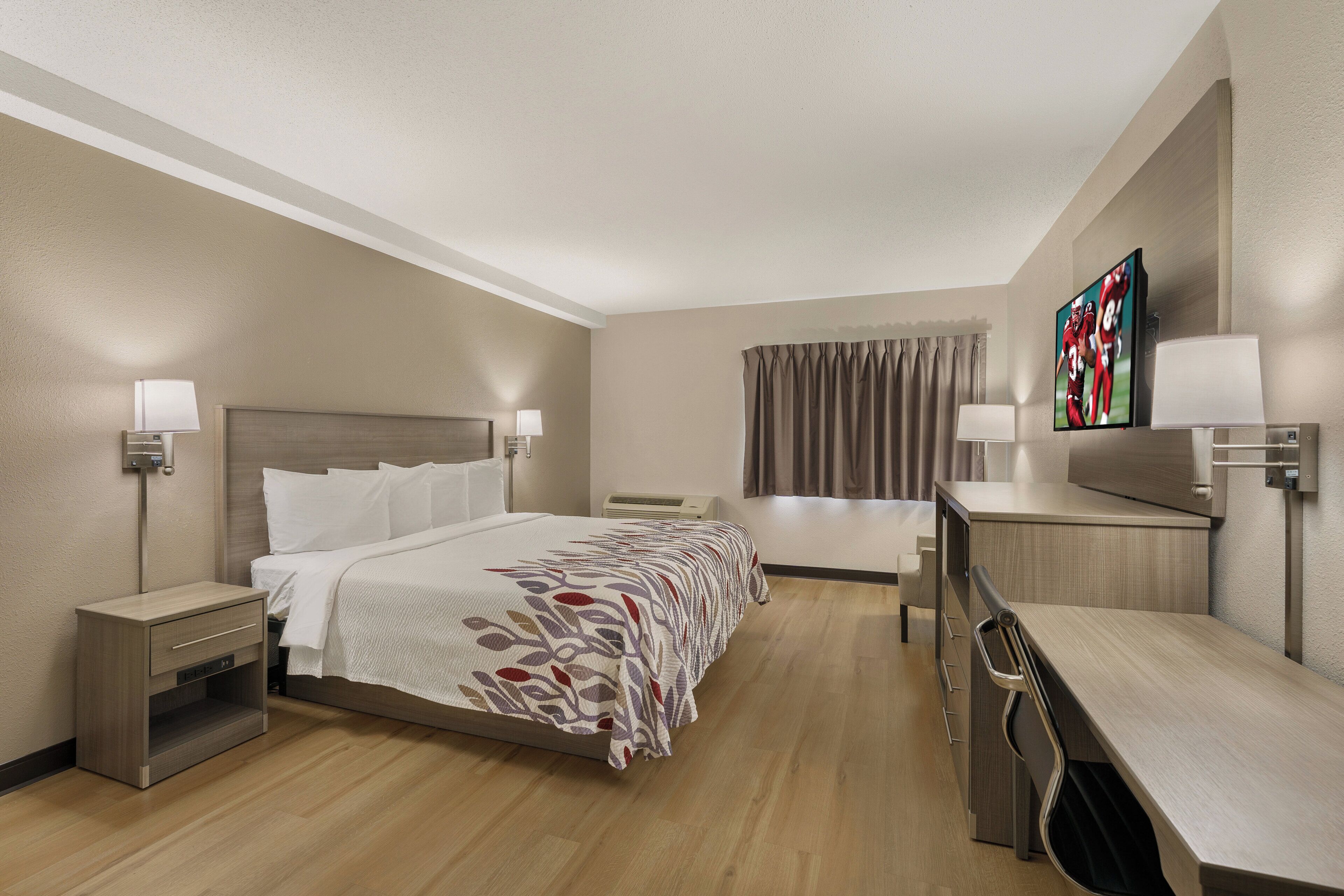 Habitación de hotel con cama, mesitas de noche, escritorio, silla, televisor y ventana con cortinas.
