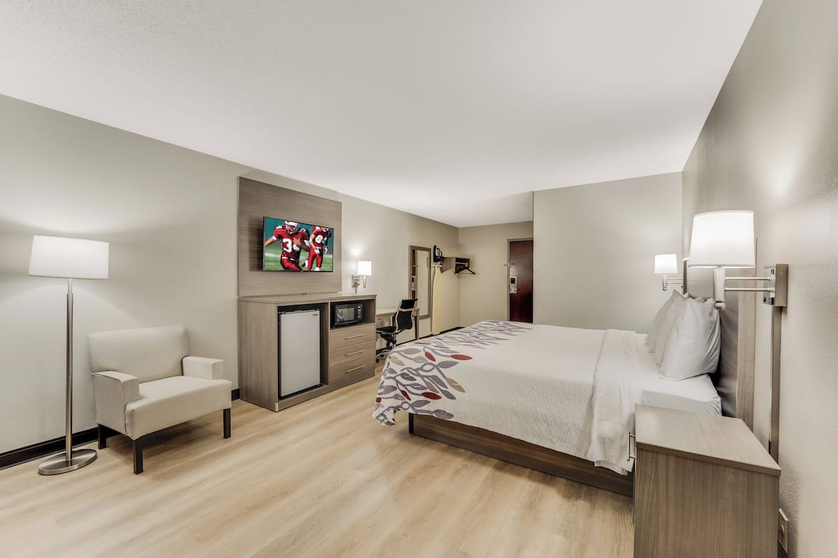 A modern hotel room with a large bed, a desk, a chair, a flat-screen TV, and a small kitchenette.