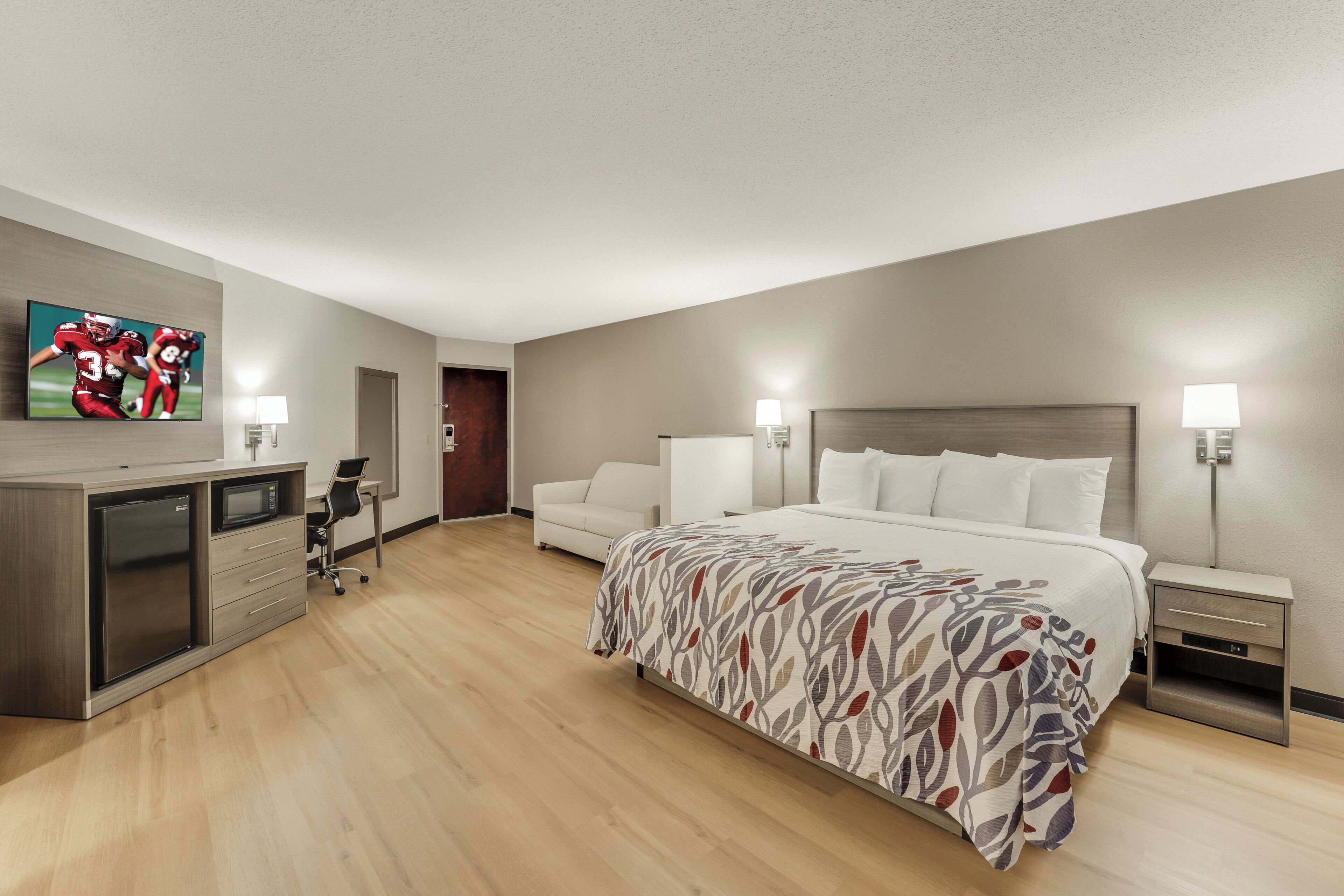 A hotel room with a bed, bedside tables, a desk with a refrigerator, a television, and a door.
