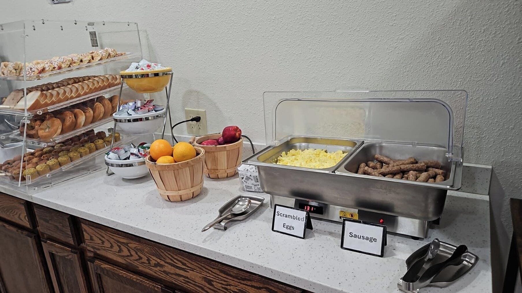 Buffet com seções identificadas para ovos mexidos, linguiça e diversos pães e doces.