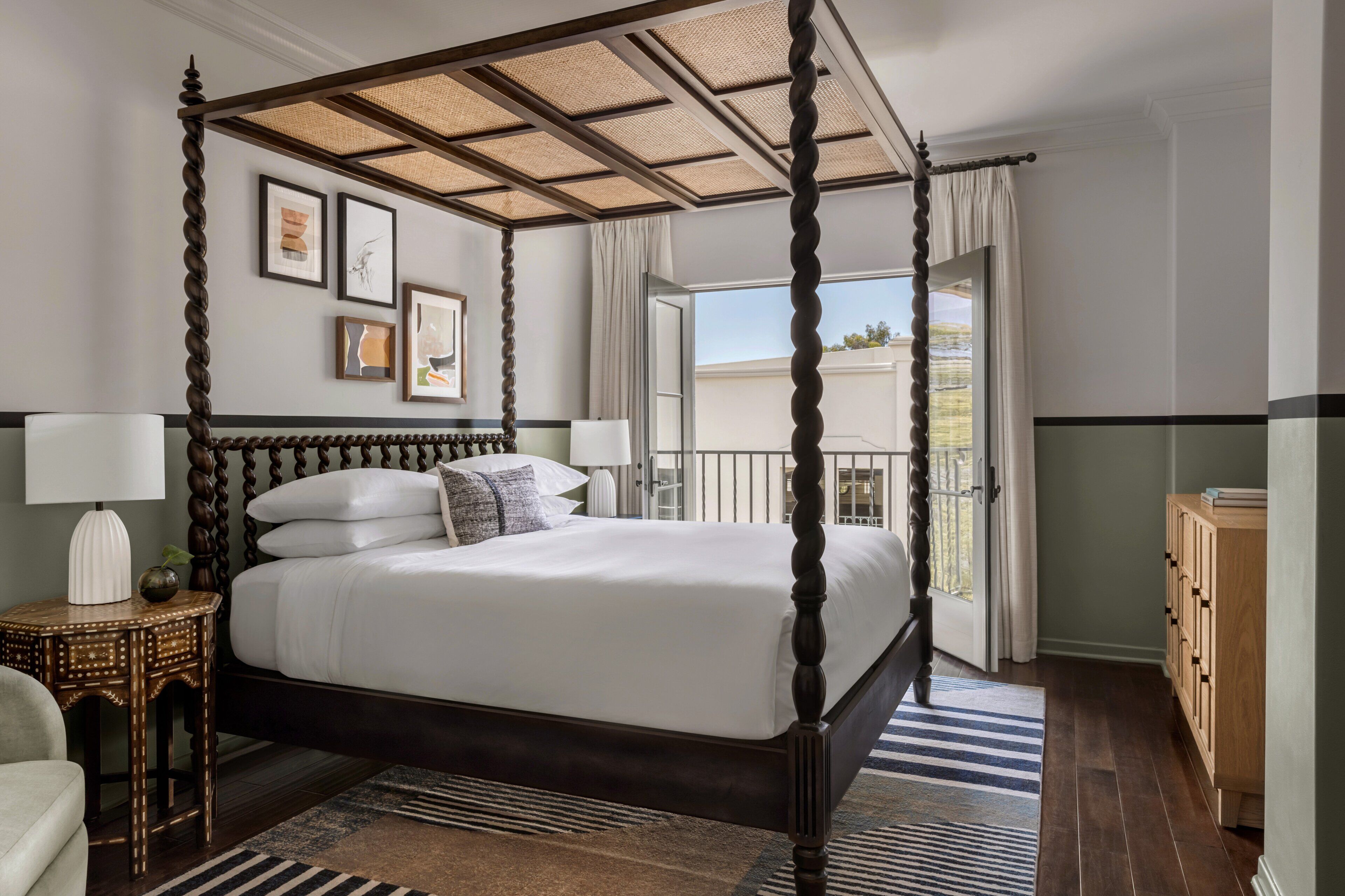 A four-poster bed with a canopy, a bedside table with a lamp, a chair, and a view of the outdoors through a window.