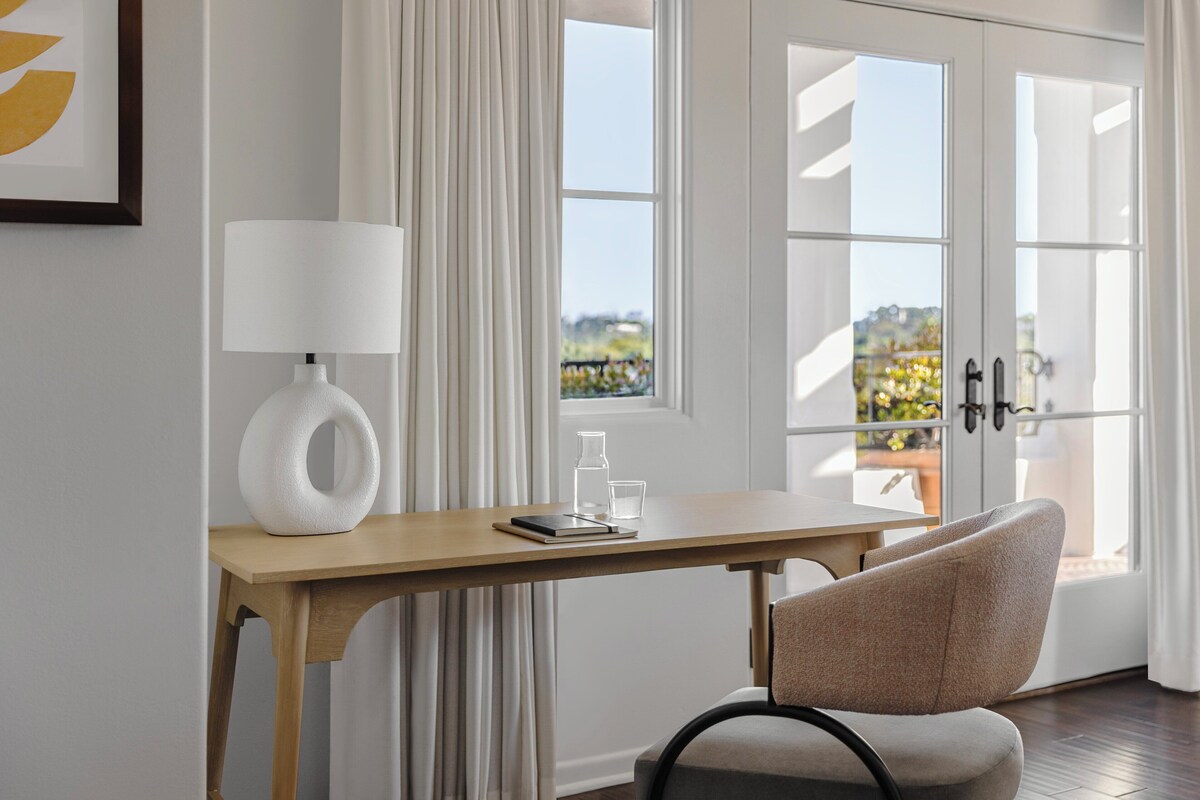 A room with a wooden desk, a chair, a lamp, and a view through glass doors.