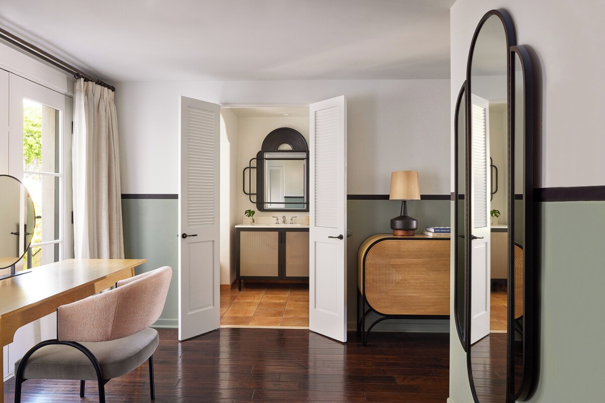 A hotel room with a wooden desk, a chair, a mirror, a dresser, and a bathroom visible through an open door.