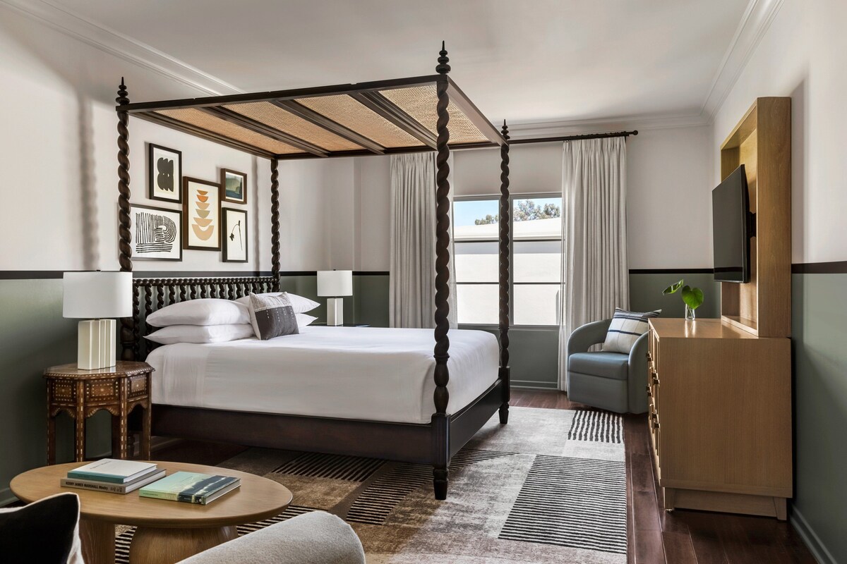 A four-poster bed with a canopy, a bedside table with a lamp, a small round table, a sofa, a wooden dresser, and framed artwork on the wall.