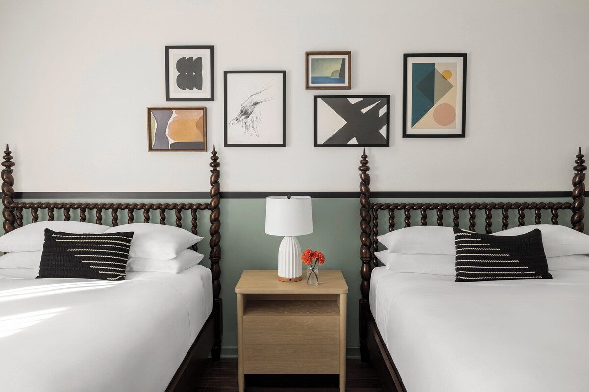 A hotel room with two beds, a nightstand with a lamp, and four framed artworks on the wall.