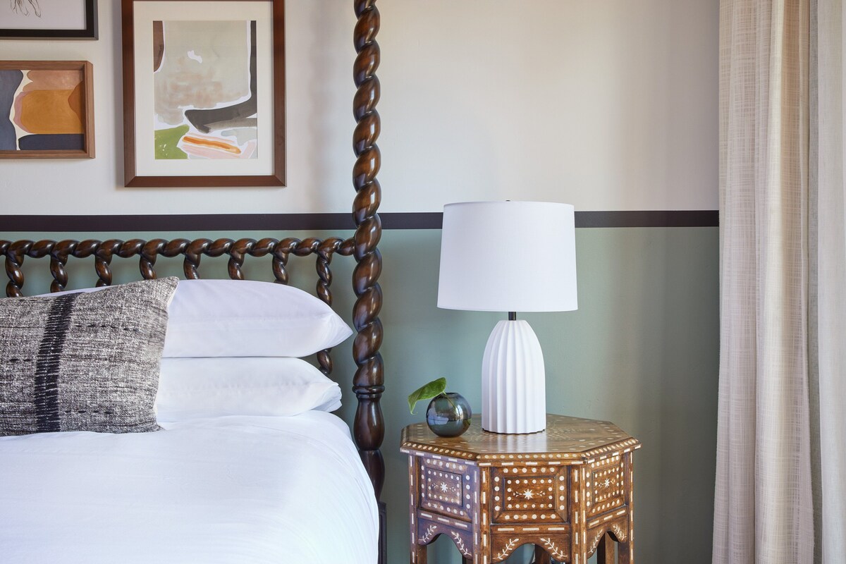 A neatly made bed with white linens and a gray patterned pillow, a wooden bedside table with a white lamp, and two framed abstract paintings on the wall.