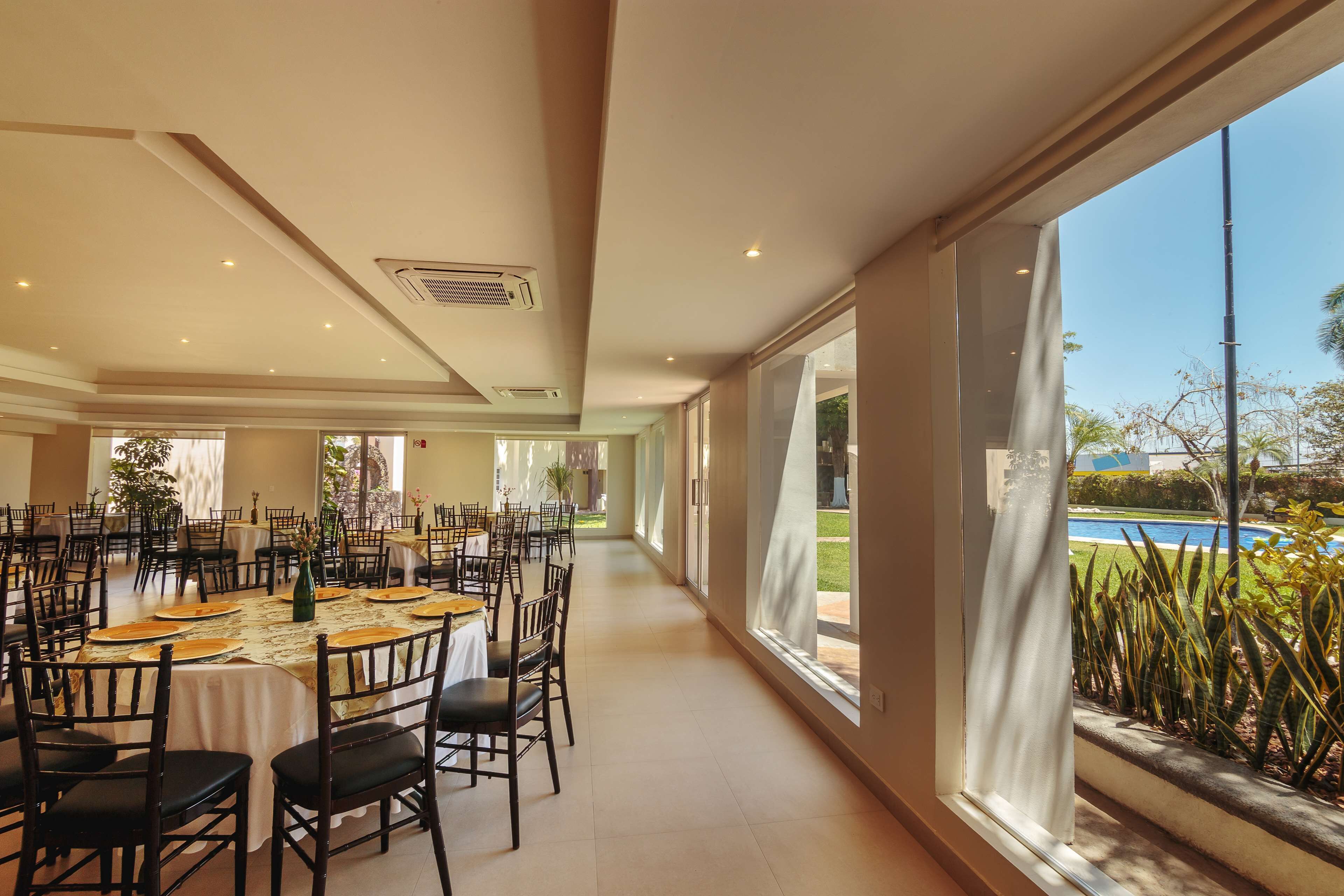 A spacious dining area with multiple tables and chairs, large windows with curtains, and a view of a swimming pool.