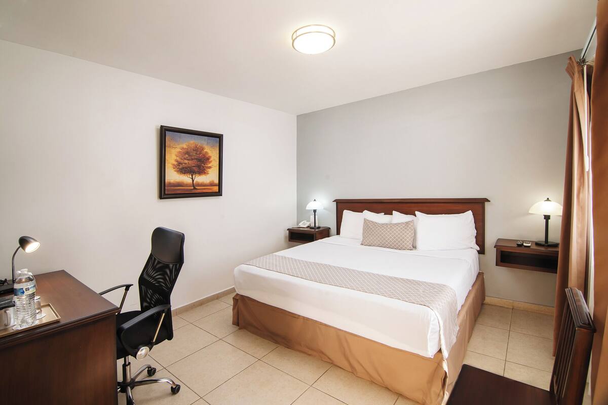 Ein Hotelzimmer mit Bett, Schreibtisch, Stuhl, Nachttischen, Lampen und einem Bild an der Wand.
