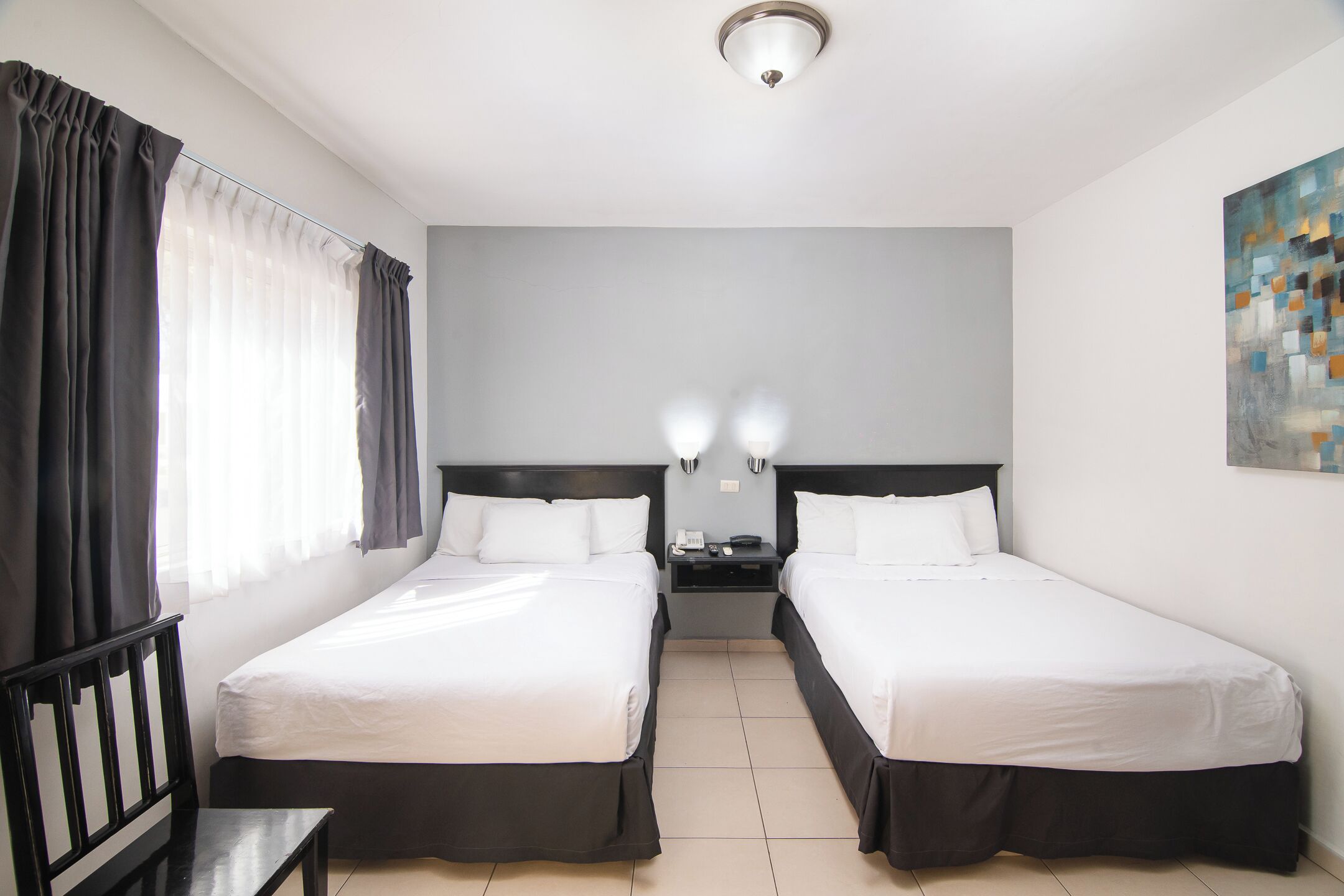 A hotel room with two single beds, a small table, a bench, a nightstand with a phone and lamp, and a painting on the wall.