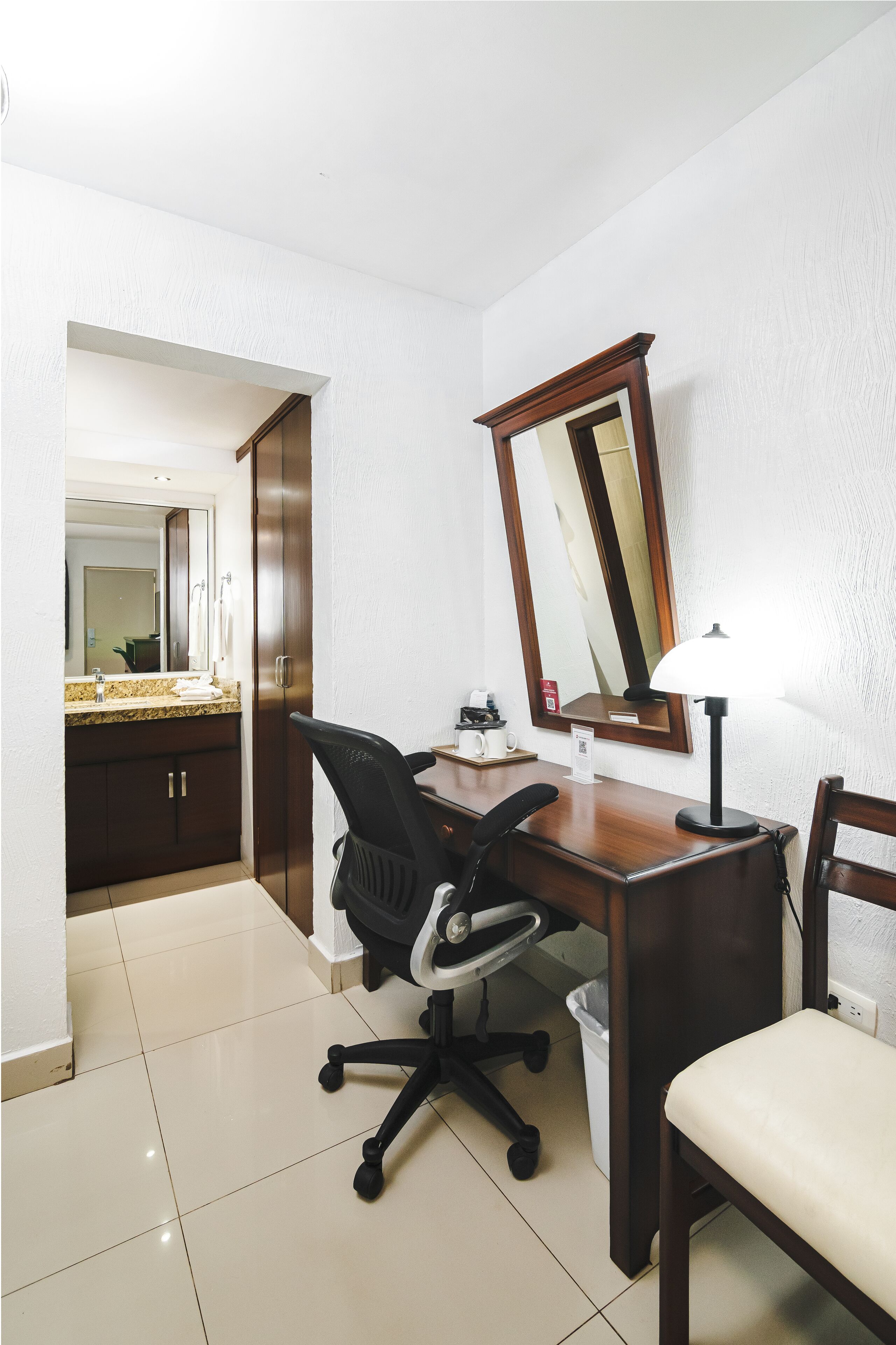 Habitación de hotel con un escritorio de madera, una silla, una lámpara, un espejo y un baño visible a través de una puerta abierta.
