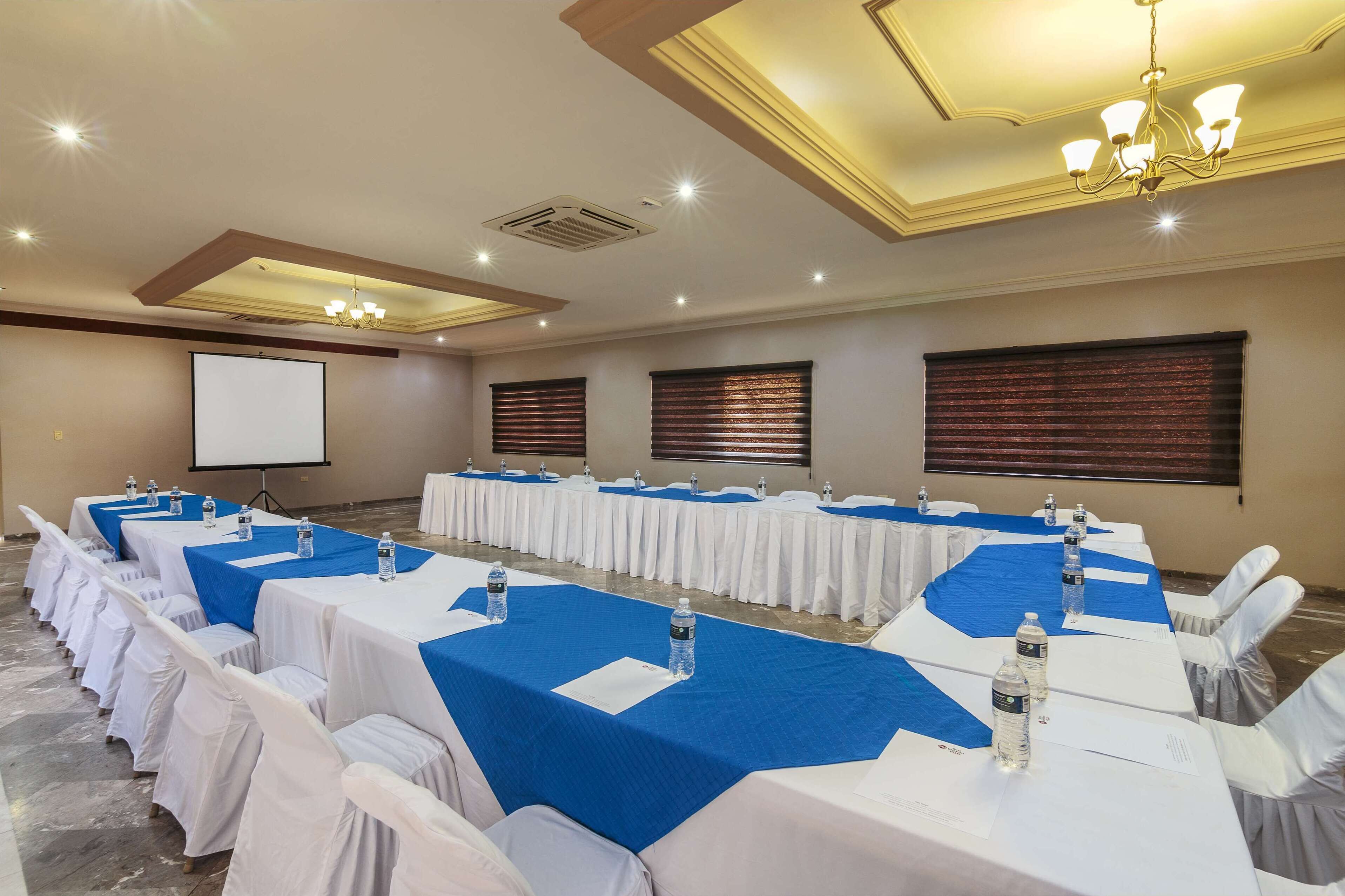 Un salón de conferencias con mesas largas, manteles blancos y azules, y botellas de agua.