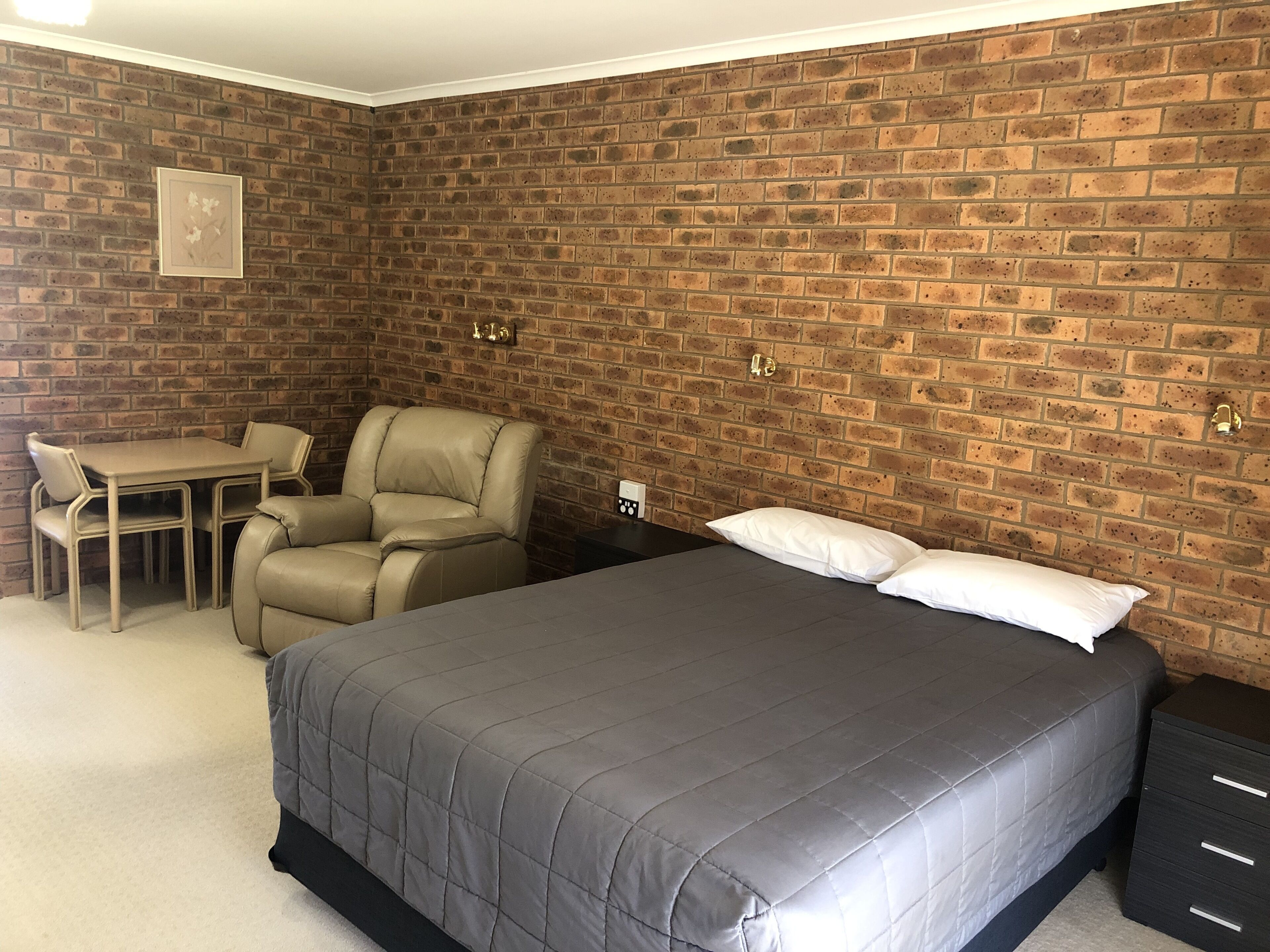 A room with a brick wall, a bed with a grey quilt, a chair, a small table, and a nightstand with drawers.