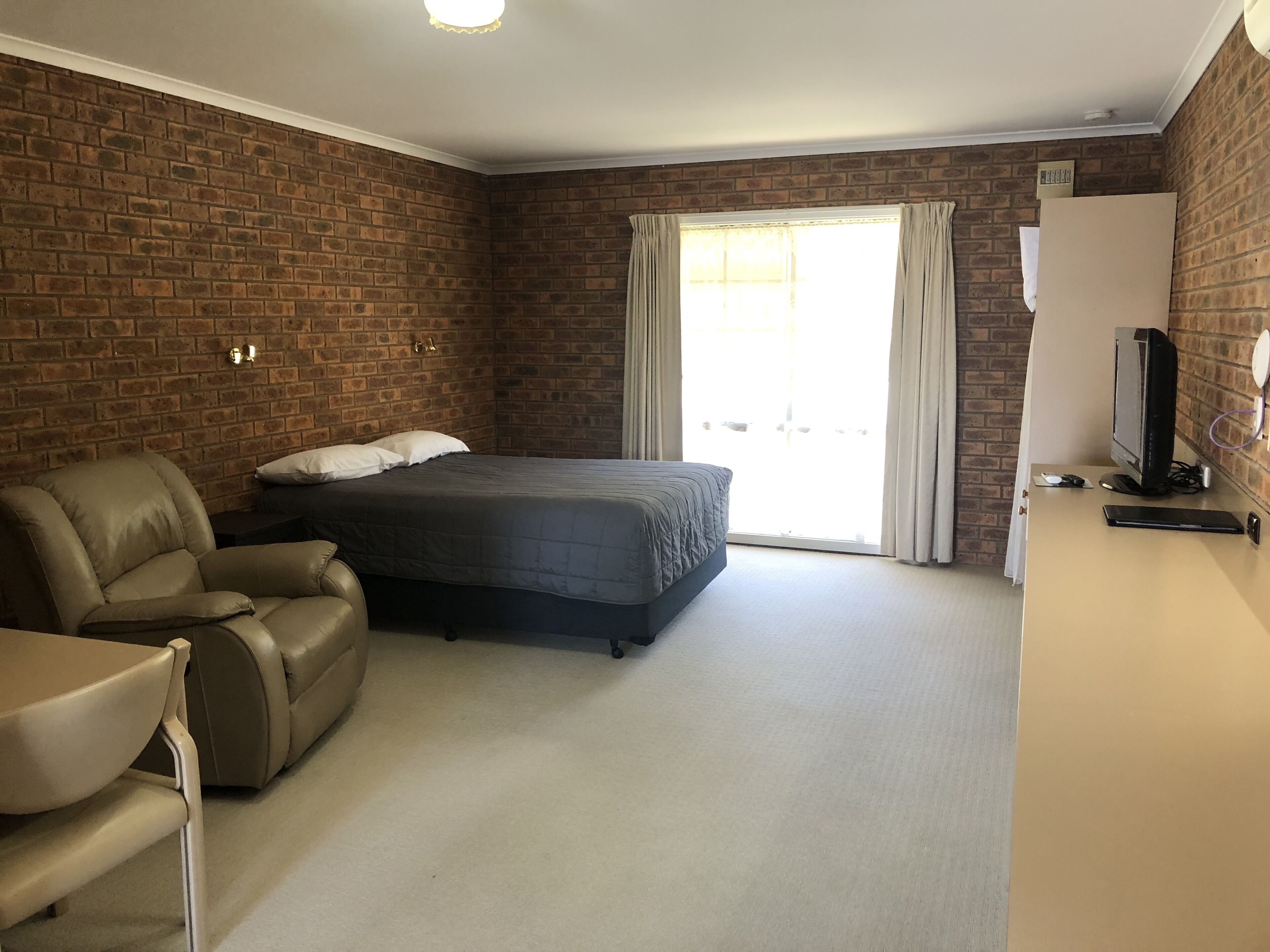 A bedroom with a bed, a leather chair, a desk with a computer, and a brick wall.