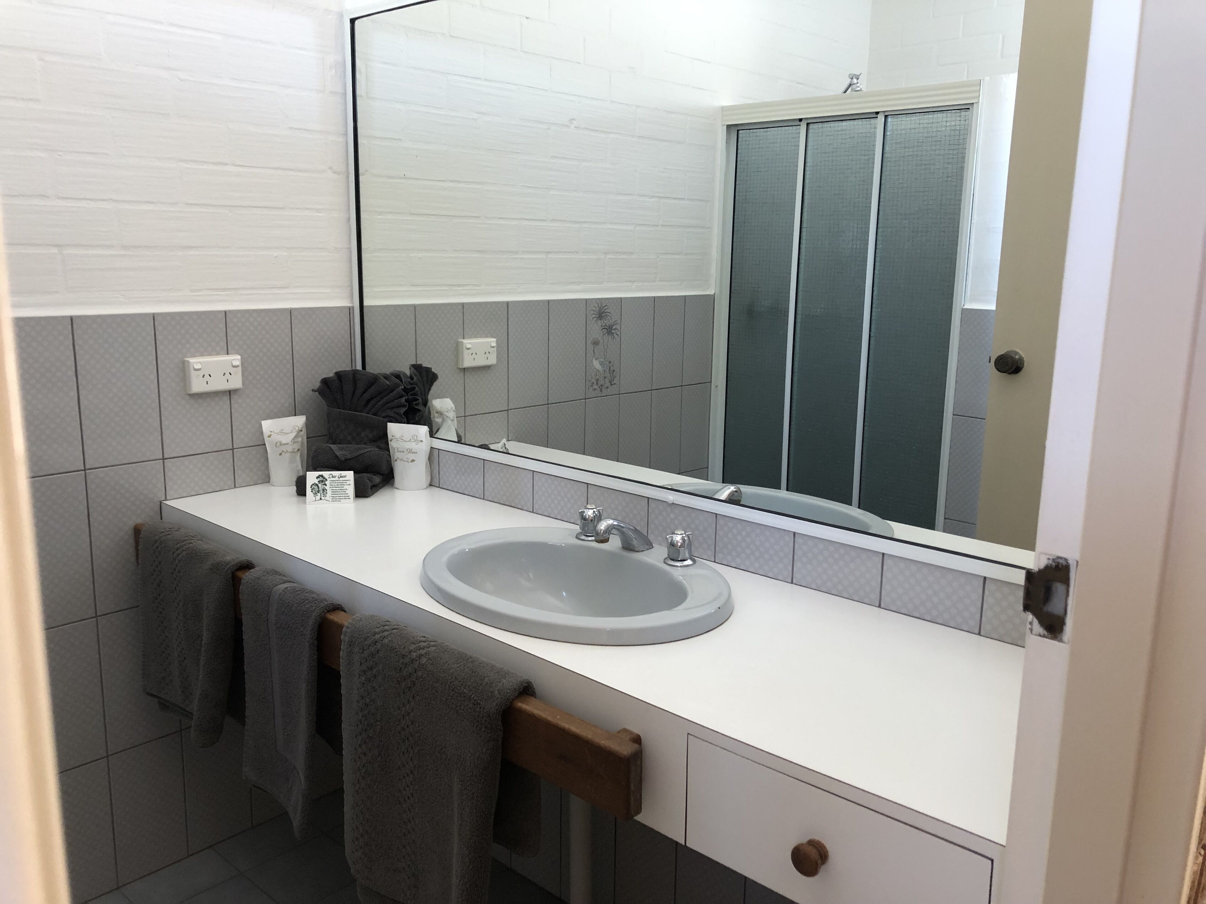Baño con lavamanos blanco, espejo y paredes de azulejos. Hay toallas colgadas en un perchero y un estante pequeño con artículos de aseo.
