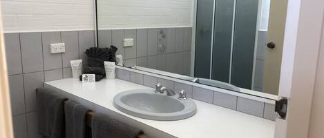 Baño con lavamanos blanco, espejo y paredes de azulejos. Hay toallas colgadas en un perchero y un estante pequeño con artículos de aseo.