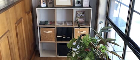 Un área de recepción con un mostrador de madera, una planta en una maceta negra y un estante blanco con varios objetos.
