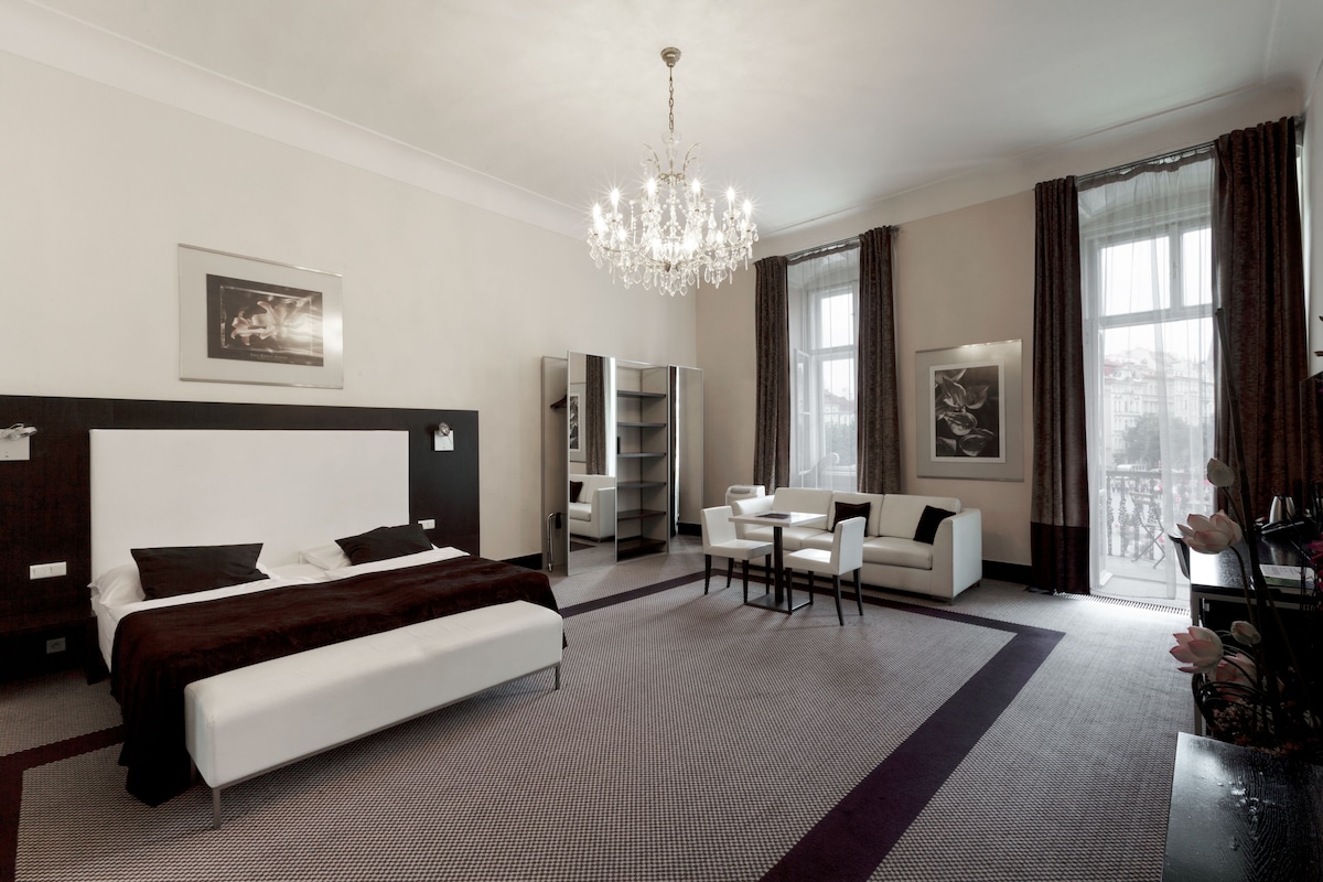 Een moderne hotelkamer met een groot bed, een zithoek met een bank en stoelen, een kroonluchter en grote ramen met gordijnen.