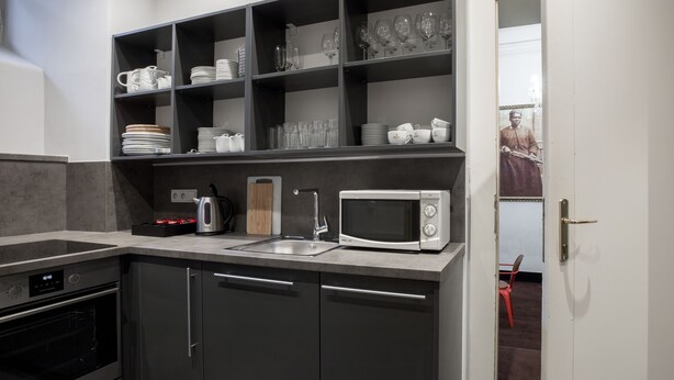 A modern kitchen with a built-in microwave, a kettle, and a sink. There are shelves above the counter holding plates and glasses.