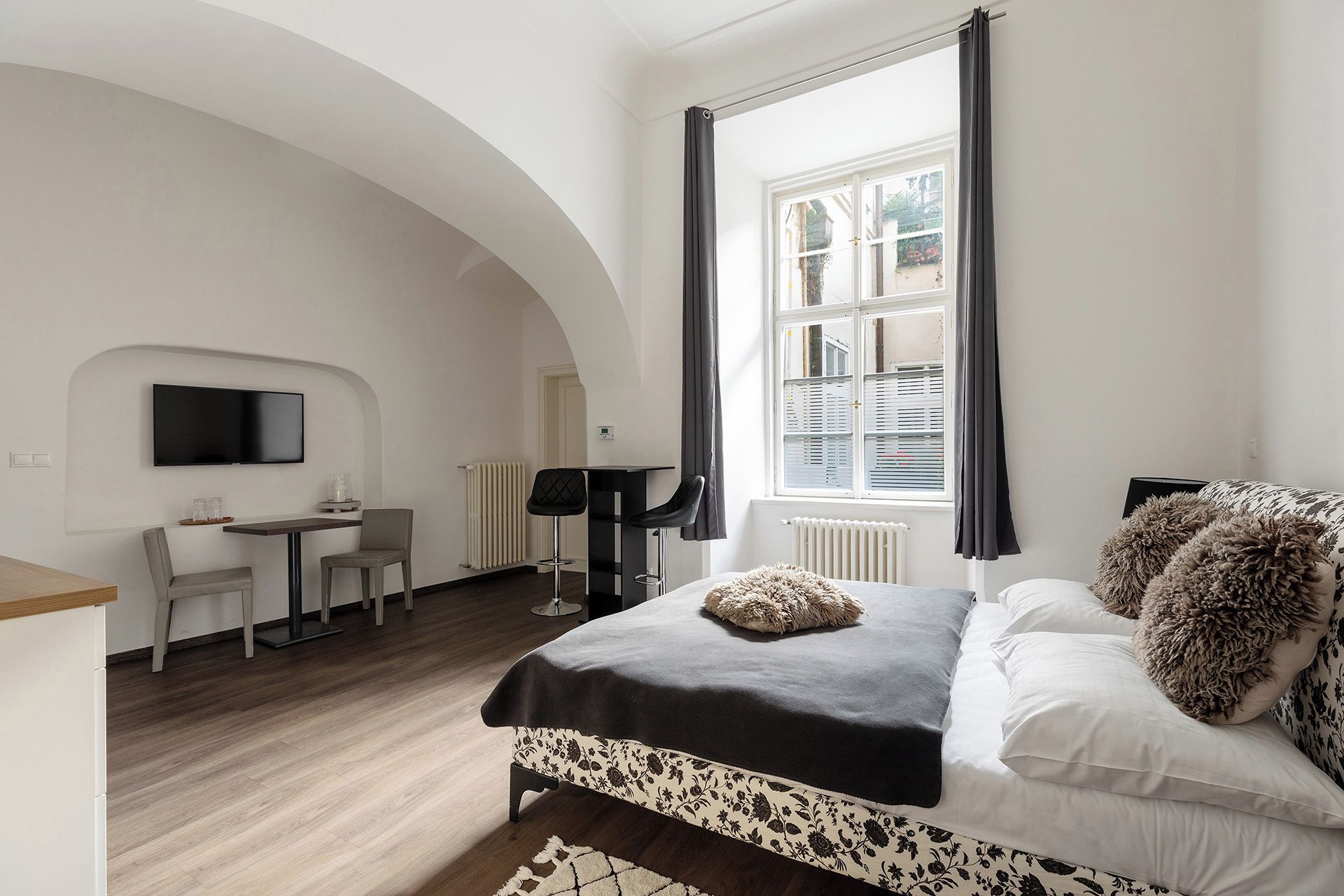 A bedroom with a bed, a desk, a chair, a TV, and a window with curtains.