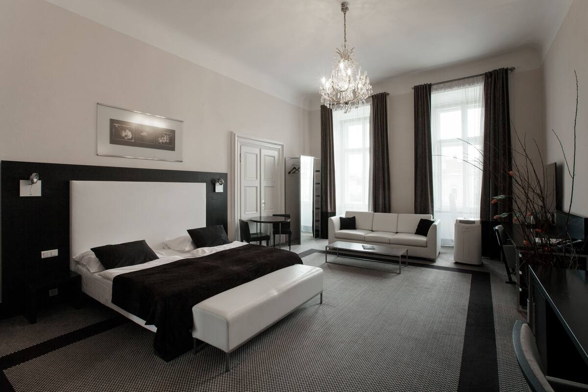 A modern hotel room with a large bed, a white sofa, a small table, and a chandelier.