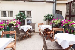 An outdoor patio with wicker furniture and white tablecloths, surrounded by potted plants with pink and white flowers.