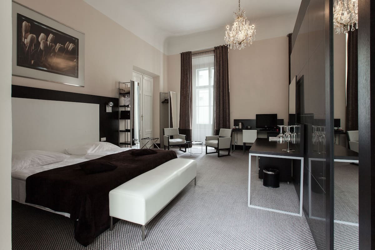 A modern hotel room with a large bed, a desk, a chair, a bench, a TV, and a chandelier.