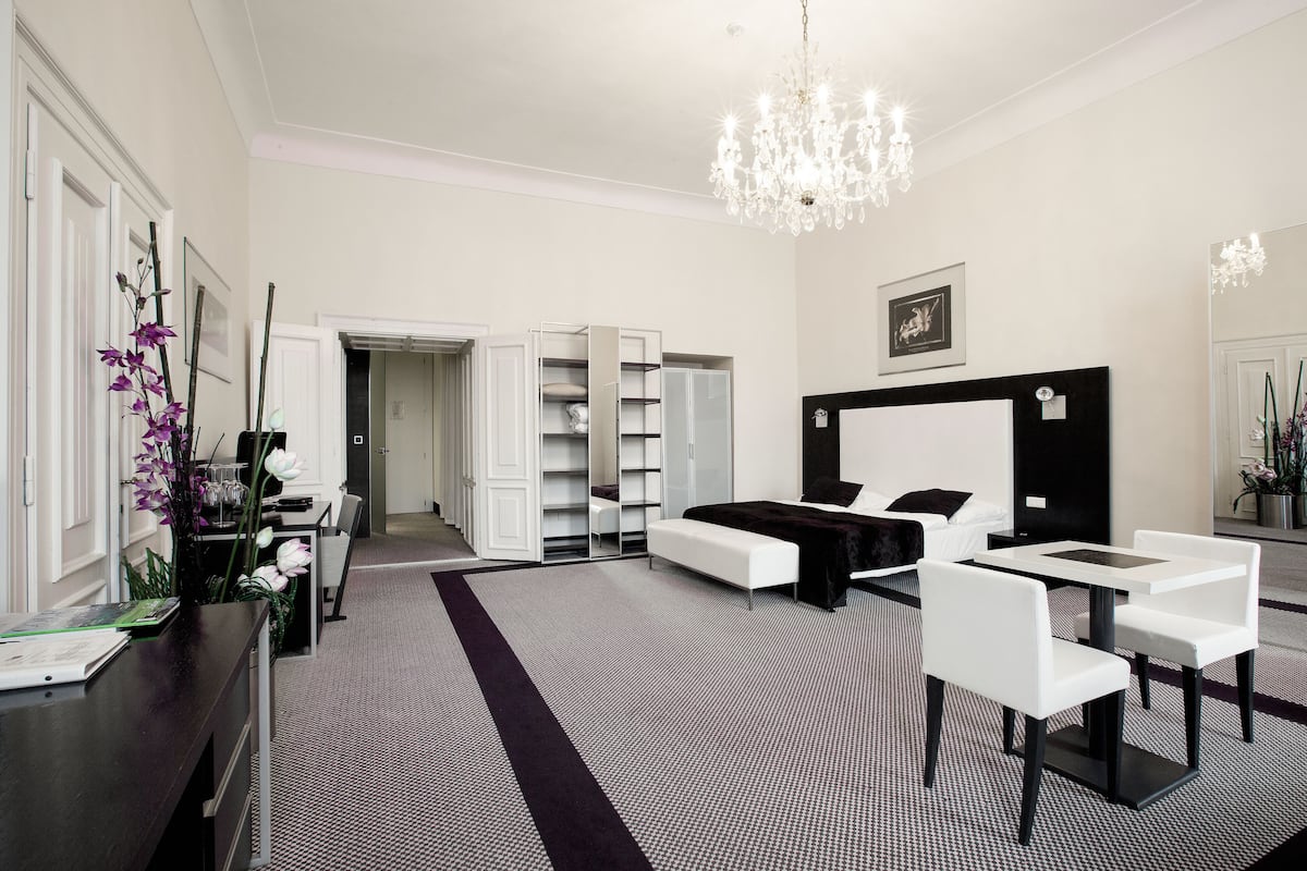 A modern bedroom with a large bed, bedside tables, a desk, and a chair. There is a wardrobe with a mirror and a chandelier.