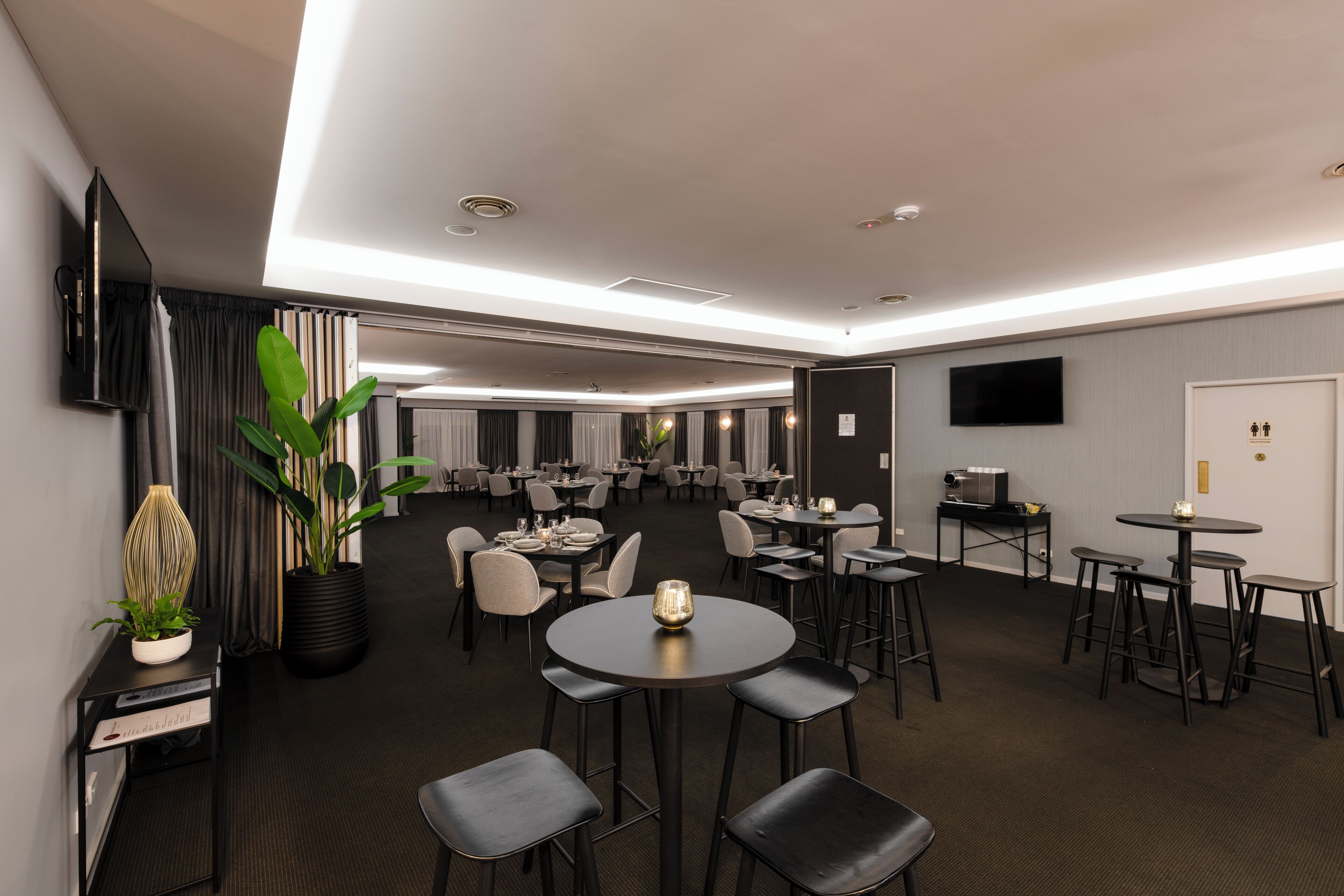 A modern hotel lobby with multiple dining areas, a reception desk, and a flat-screen TV mounted on the wall.