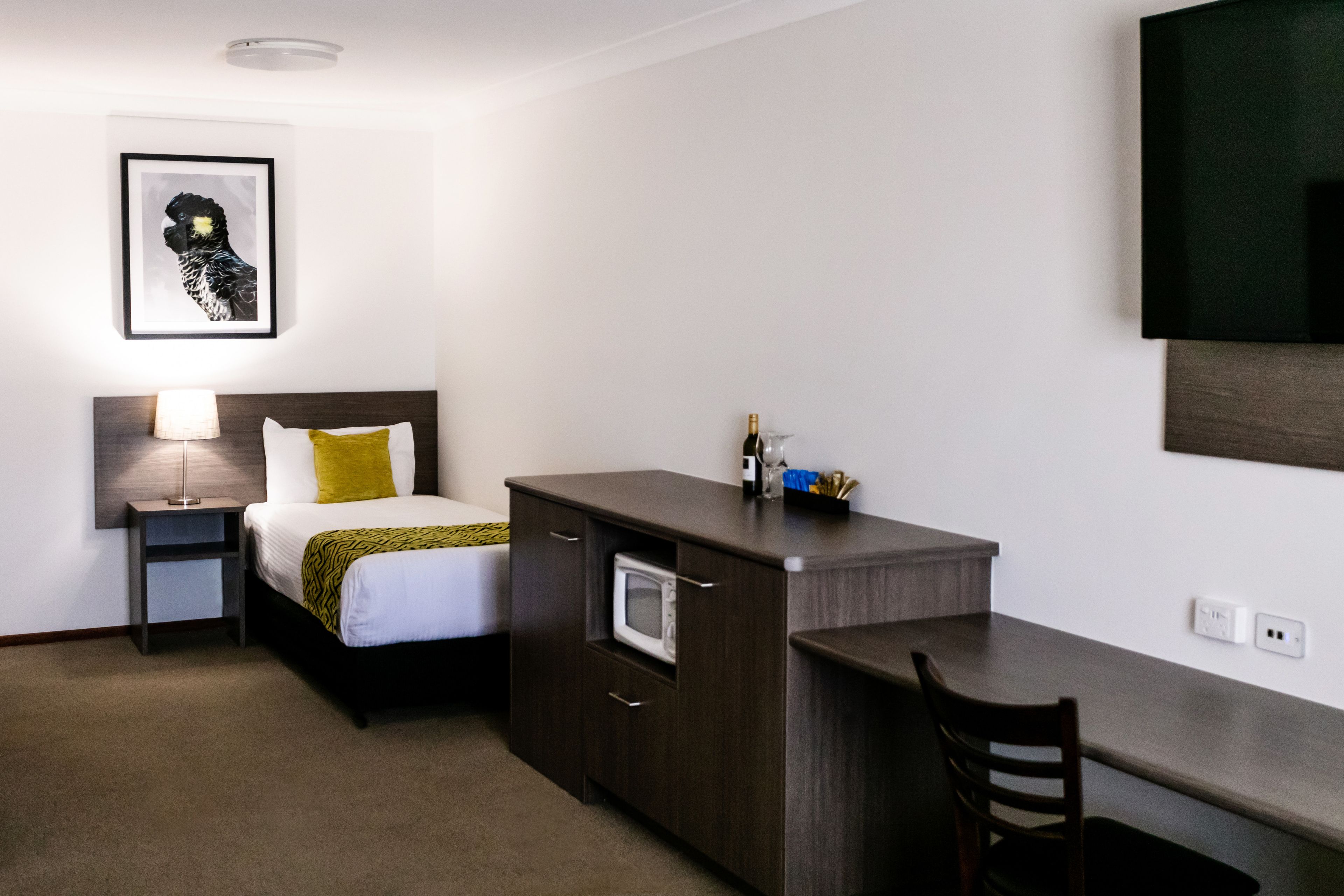 A hotel room with a bed, bedside table, lamp, microwave, and a desk with a chair.
