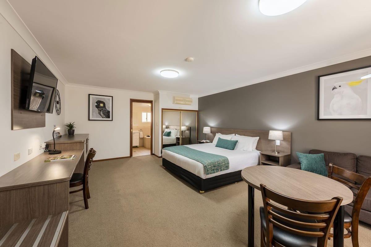 A hotel room with a bed, a desk, a chair, a table, a TV, and a bathroom visible through an open door.