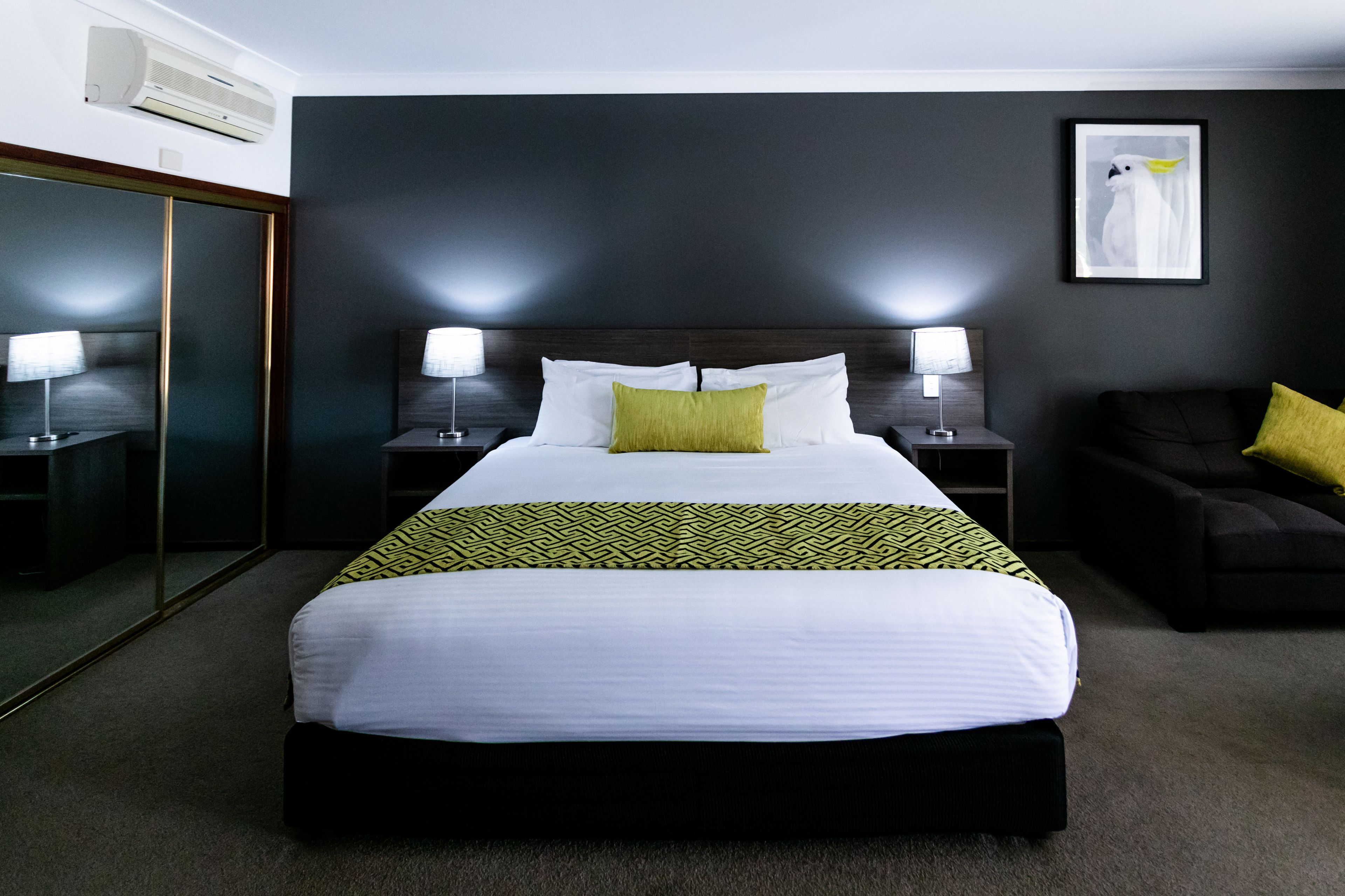 A hotel room with a large bed, two bedside tables with lamps, a dark-colored sofa, and a framed picture on the wall.