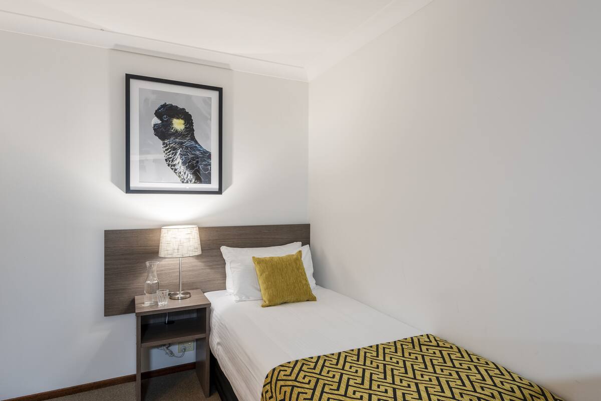 A neatly arranged hotel room with a bed, a bedside table with a lamp, a framed artwork on the wall, and a patterned throw blanket.