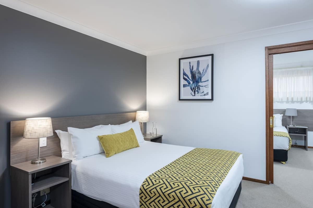 A hotel room with a bed, bedside lamps, a nightstand, a framed artwork, and a door leading to another room.