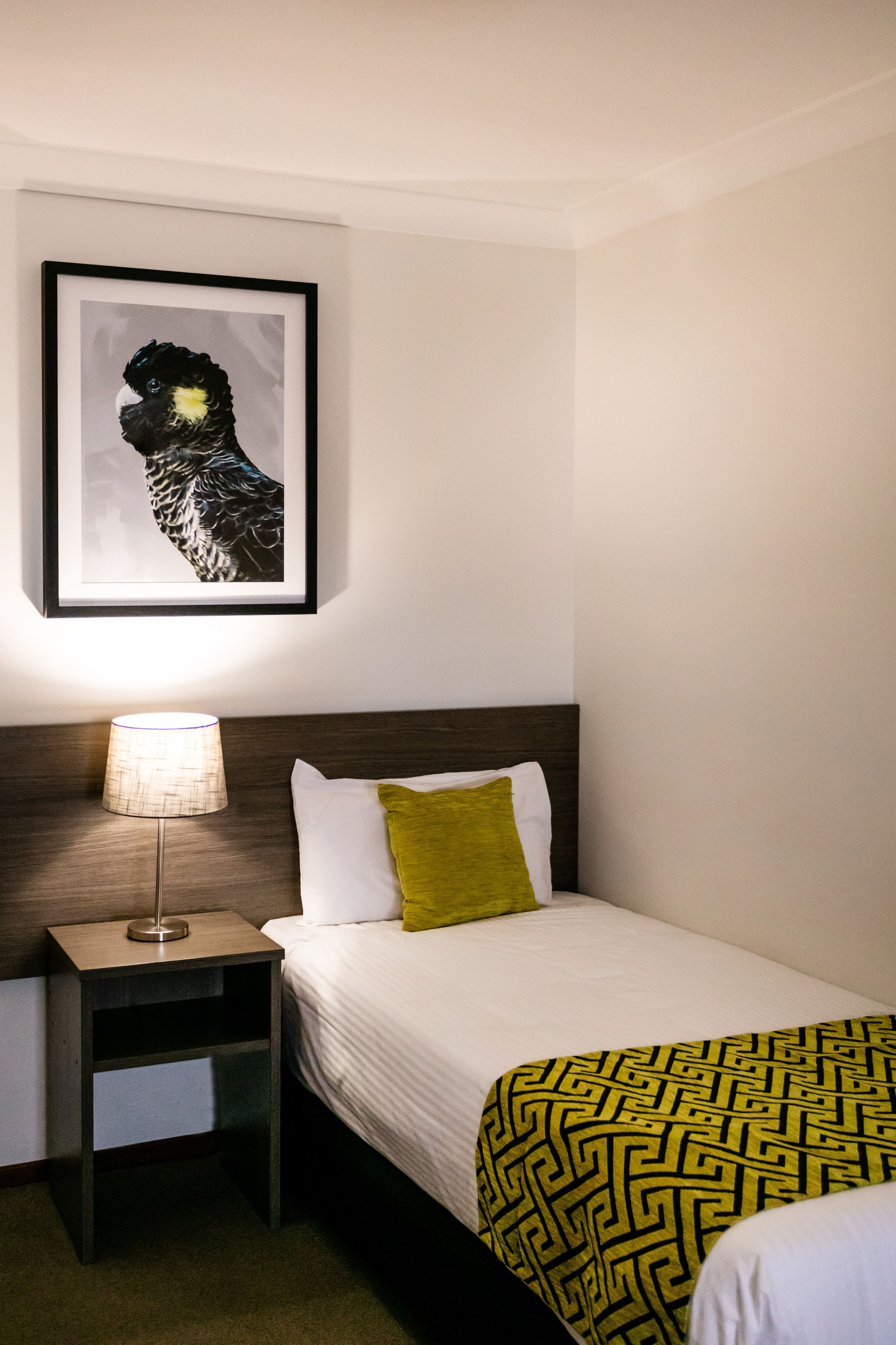 Ett hotellrum med en säng, en sänglampa, ett inramat konstverk på väggen och en mönstrad pläd.