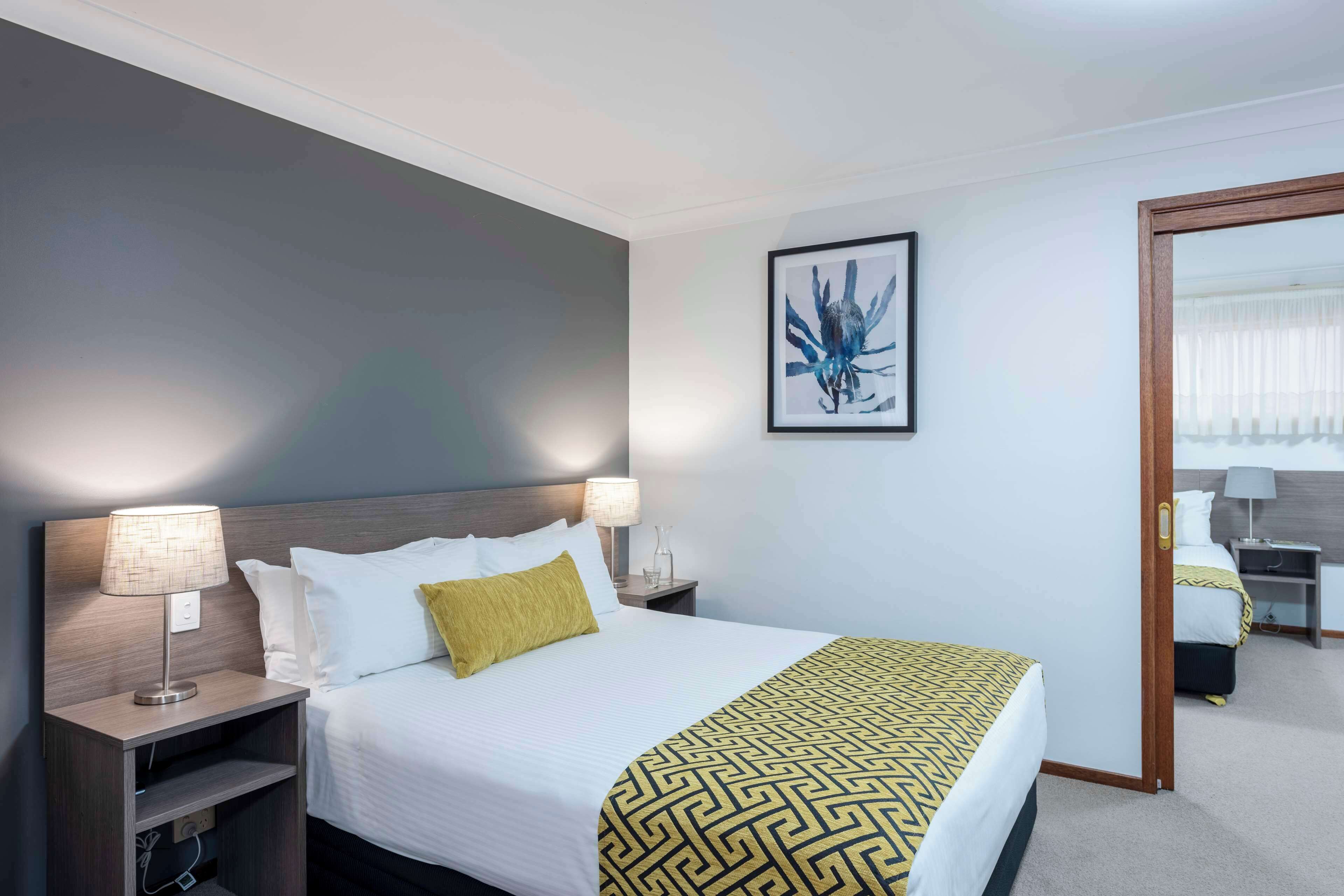 Habitación de hotel con cama, dos mesitas de noche con lámparas, una obra de arte enmarcada en la pared y una puerta que da a otra habitación.