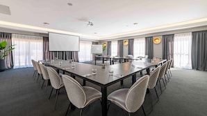 Un salón de conferencias con una larga mesa, sillas, una pantalla de proyector y una planta grande en maceta.