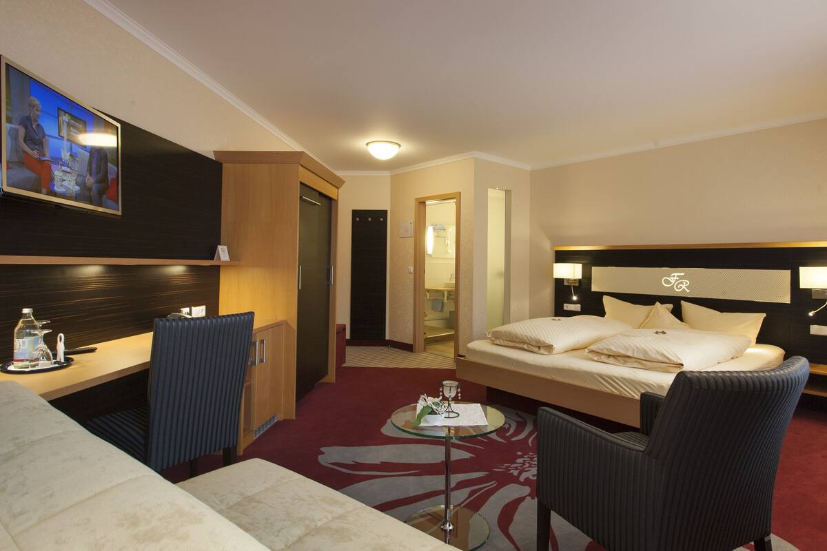 Habitación de hotel con cama, escritorio, silla, televisión y baño visible a través de una puerta abierta.