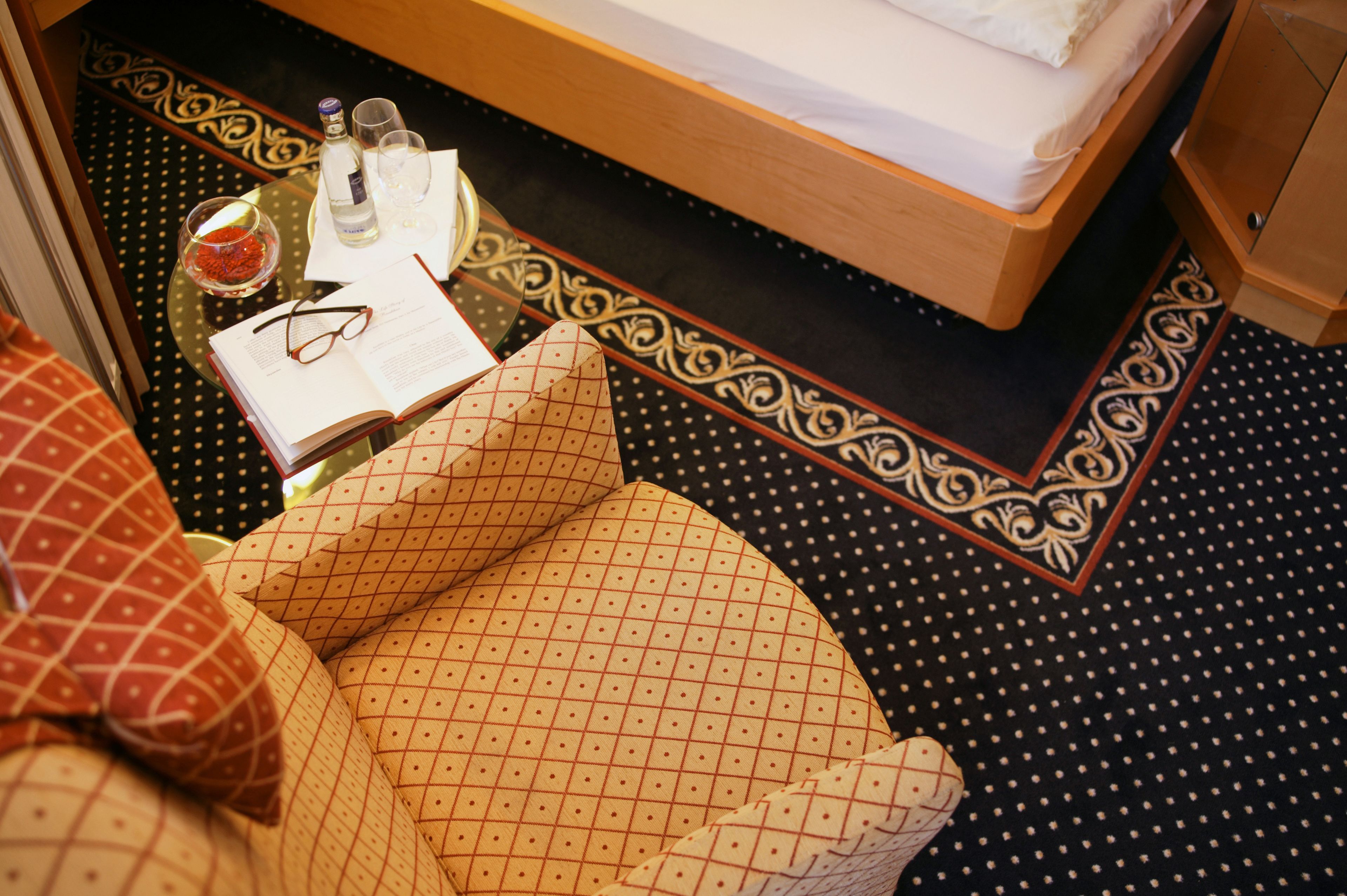 Une chambre d’hôtel bien rangée, avec un lit, une petite table sur laquelle se trouvent un livre, des lunettes et une bouteille.