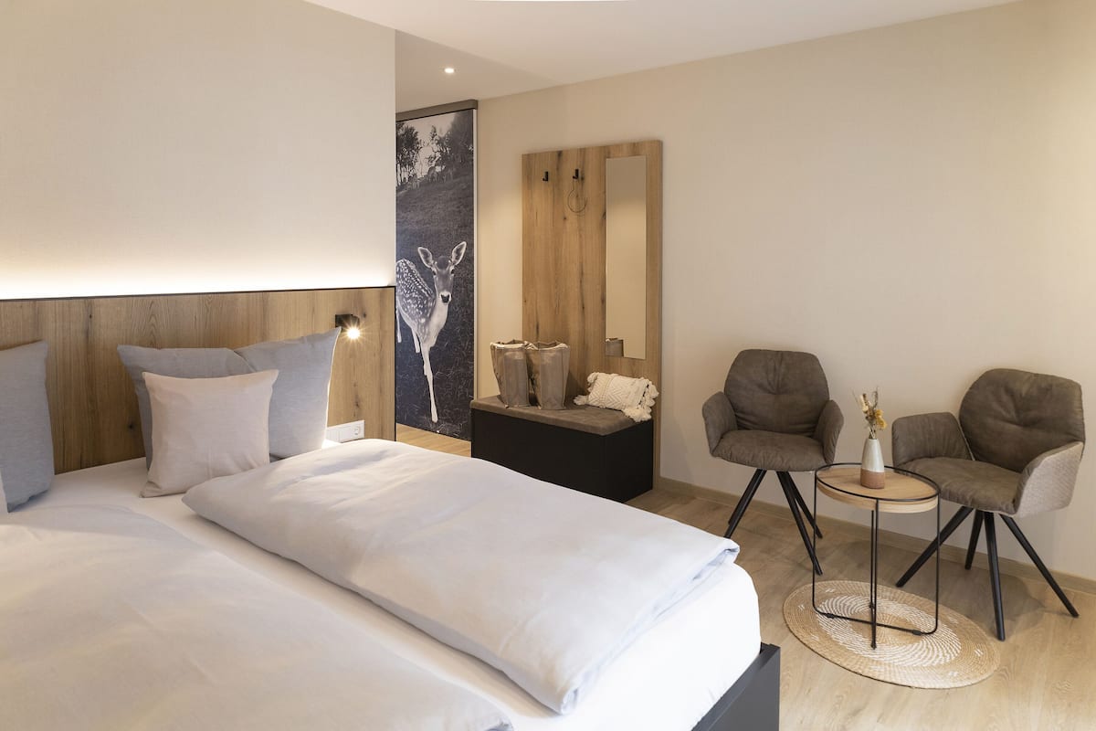 Una habitación de hotel moderna con una cama grande, dos sillones, una pequeña mesa redonda, un espejo y una obra de arte colgada en la pared.