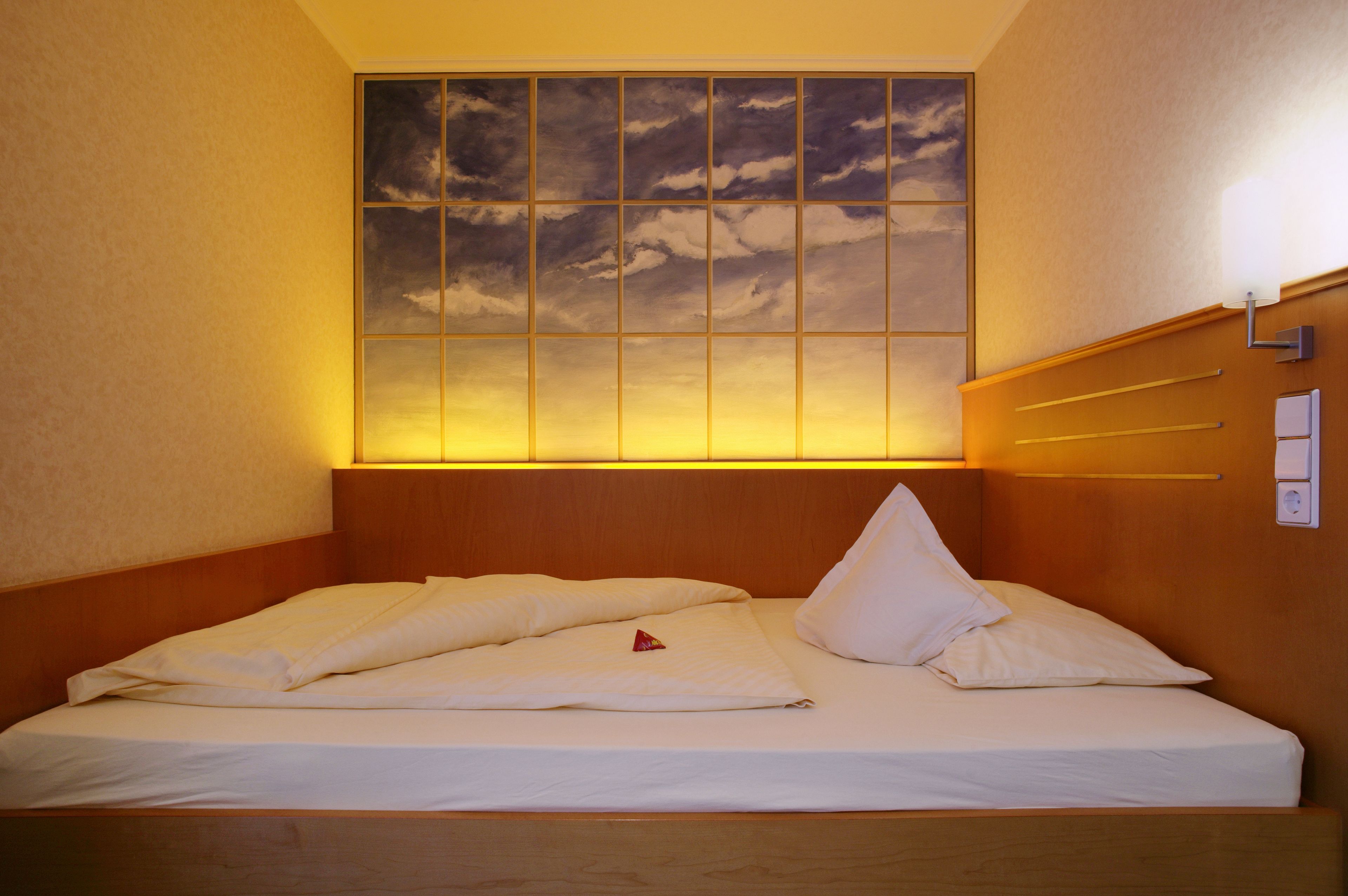 Ein Hotelzimmer mit Bett, Kopfteil, Wandlampe und einem großen Fenster mit Blick auf Wolken.