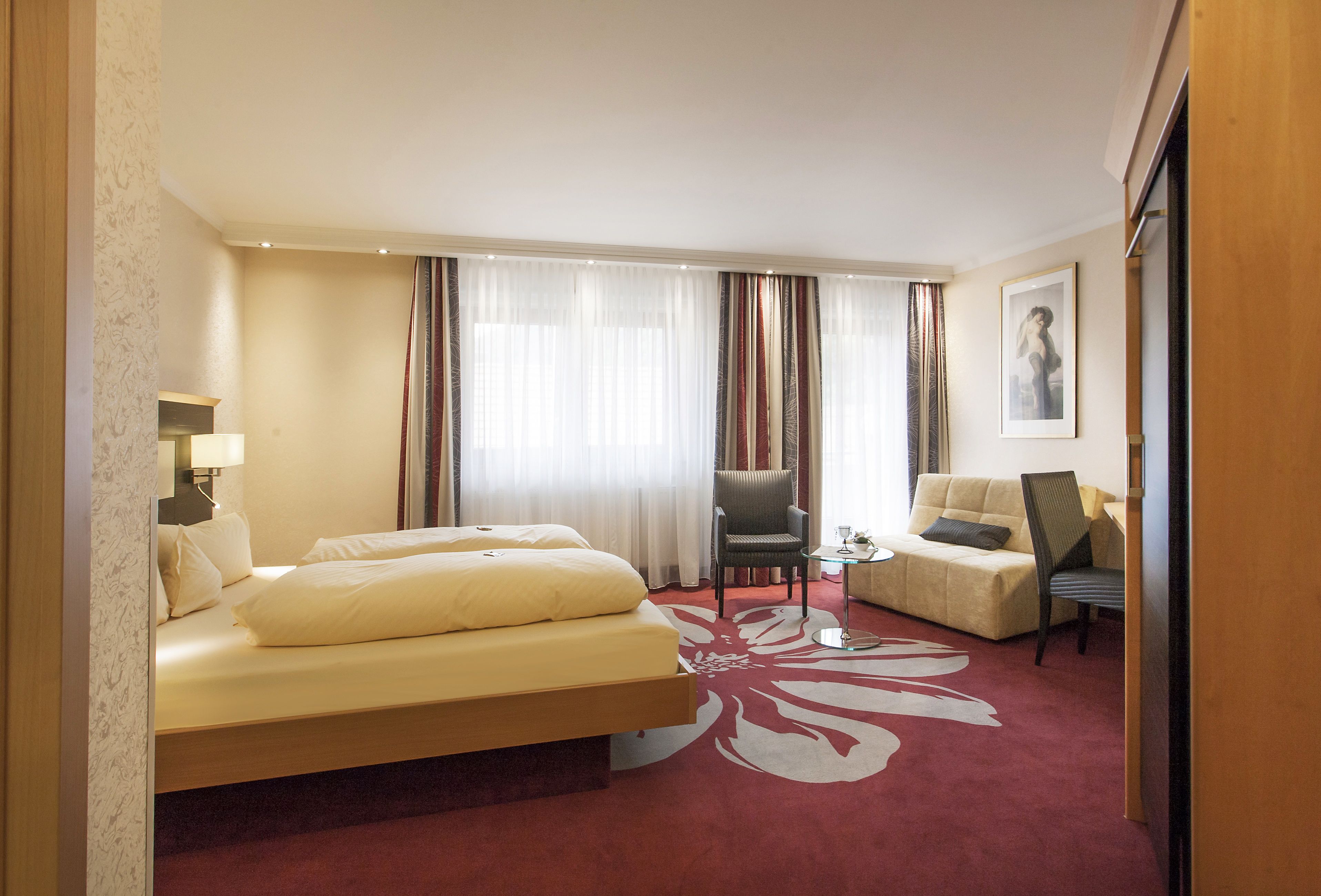 A hotel room with a bed, two chairs, a small table, and a large window with curtains.