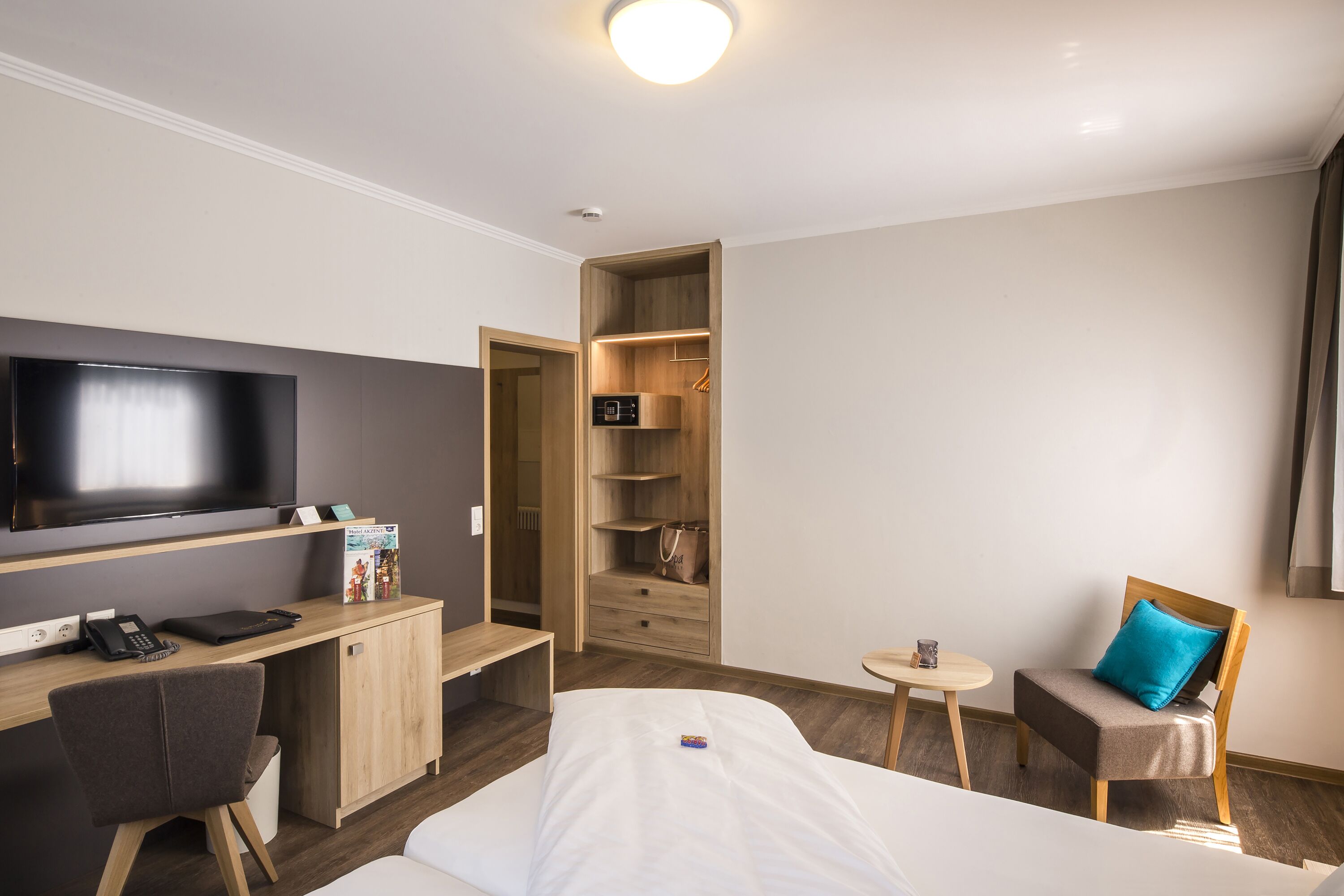 A hotel room with a bed, a desk with a computer, a chair, a TV, a wardrobe, and a small table with a cup.