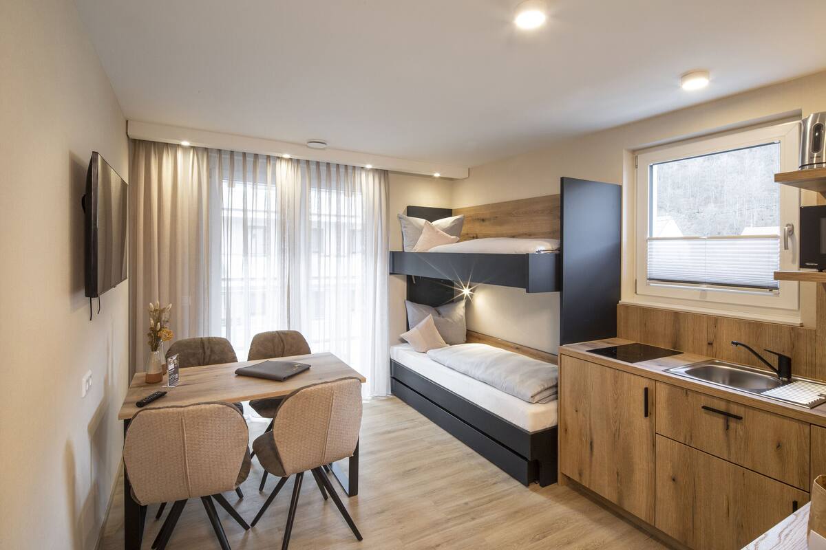 Habitación de hotel compacta con literas, kitchenette, comedor y televisor montado en la pared.