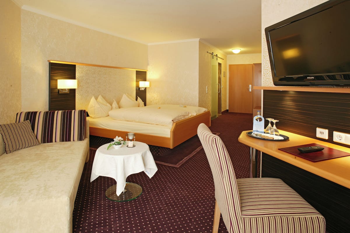 Habitación de hotel con cama, sofá, una mesita con un florero, una silla y televisor.