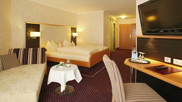 A hotel room with a bed, a sofa, a small table with a vase, a chair, and a TV.