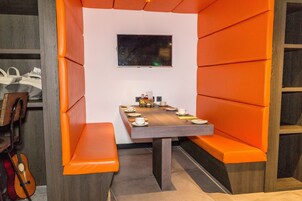 Un comedor con mesa de madera dispuesta para cuatro personas, asientos con cojines anaranjados, un televisor de pantalla plana en la pared y una guitarra apoyada contra la pared.