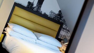 A bed with white linens and a yellow headboard, a small round table, and a wall with a black and white cityscape mural.