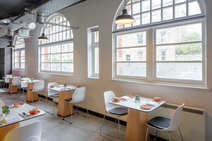 Un comedor moderno con mesas y sillas, ventanas amplias con vistas a edificios, e iluminación colgante.